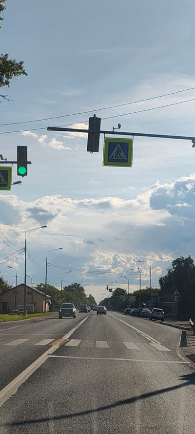 The image depicts a street scene in Неёлово-1, Russia, under a partly cloudy sky. In the foreground, a two-lane asphalt road is visible, marked with white lines. A pedestrian crossing, indicated by white stripes, is present on the road. Several vehicles are driving on the road, including a dark SUV in the center lane, a white car behind it, and other cars further in the distance. On the left side of the road, a low building with a brown facade and a pitched roof is situated, with trees behind it. Streetlights are visible along both sides of the road.

Overhead, a traffic light system is mounted on a horizontal pole crossing the road. The traffic light on the left is displaying a green signal. To the right of the traffic light, a pedestrian crossing sign, a blue square with a white silhouette of a person walking, is also visible. Another identical pedestrian crossing sign is mounted further to the right on the same overhead structure. Overhead power lines are strung across the scene. In the distance, the road appears to curve slightly, with more traffic and trees lining the sides. The overall impression is of a typical suburban or semi-urban street on a bright day.