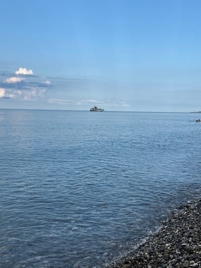 A wide shot captures a calm, blue sea under a hazy sky with scattered clouds. In the distance, a white boat with a dark roof sails on the horizon. The foreground features a pebble beach where gentle waves lap at the shore. Two individuals are visible in the water on the right side of the frame, appearing to be swimming or wading. The overall scene suggests a peaceful day at the coast in Amzara, Abkhazia.