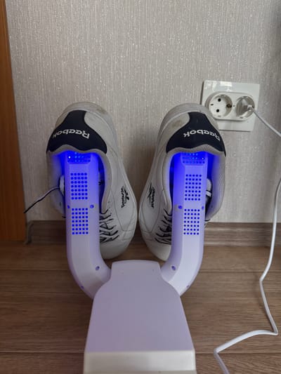 In an indoor scene, a white shoe dryer with two arms, each fitted with blue lights, is placed on a wooden floor. Two white Reebok sneakers are inserted into the arms of the dryer. A white power cord extends from the dryer to a white electrical outlet mounted on a textured wall. The outlet is visible on the right side of the image.