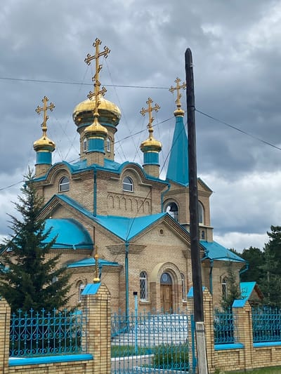 An Eastern Orthodox church with a tan brick exterior and bright blue roofs and trim is depicted under a cloudy sky. The church features multiple golden domes topped with crosses. A tall wooden utility pole stands in the foreground, partially obscuring the church. A decorative blue metal fence with brick posts encloses the church property. Evergreen trees are visible around the building and behind the fence. The location is identified as Sosnovka, Russia. There are no people visible in the image.