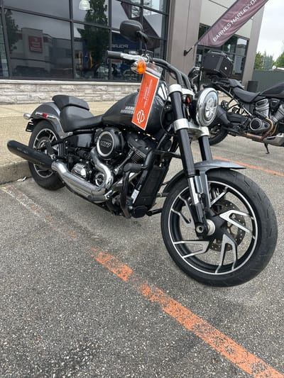 A black Harley-Davidson motorcycle is parked on an asphalt surface, next to a building with large windows. The motorcycle is the primary subject, with its front wheel and engine clearly visible. The engine has a "107" emblem on it. A price tag or information tag with the Harley-Davidson logo is attached to the handlebars. In the background, a banner with "BARNES HARLEY-DAVIDSON" is partially visible. Another motorcycle is parked behind the main subject. The scene appears to be outdoors, possibly in a dealership parking lot. Orange lines are painted on the asphalt, suggesting a designated parking area. The lighting suggests it is daytime, with overcast or cloudy weather indicated by the diffused light and lack of strong shadows. The setting is likely a commercial area in the Township of Langley, Canada. There are no people visible in the image.