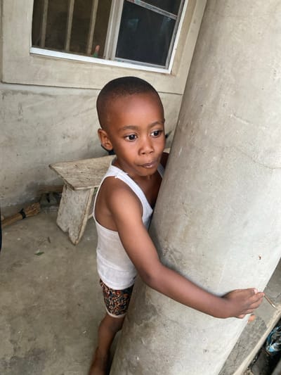 A young, dark-skinned boy with a short haircut looks into the camera, gripping a cylindrical concrete pillar. He wears a white tank top and printed shorts. The scene includes a window frame with a white edge and gray concrete around it. A wooden pallet and a broom rest on the gray concrete floor. The composition is slightly angled, giving a dynamic perspective.