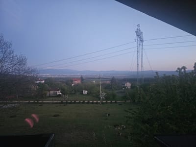 The image shows a landscape in Gradets, Bulgaria at dusk or dawn. In the foreground is a grassy yard, with trees on either side, partly obscuring the view. A few houses are visible in the middle ground, interspersed with trees and shrubbery. In the background is a panoramic view of a city with snow-capped mountains in the far distance. An electrical transmission tower stands prominently on the right side of the image. Power lines stretch across the scene. The sky is a light blue, indicating twilight. There are some blurred red lights (possibly camera artifacts) visible in the lower left portion of the yard. A dark roof edge is seen at the bottom.
