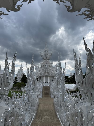The image shows the Wat Rong Khun, commonly known as the White Temple, in the city of Ban Rong Khun, Thailand. The temple is an ornate Buddhist temple that features intricate white architecture adorned with reflective mosaic pieces. In the foreground, a pathway leads towards the temple entrance, flanked by decorative white structures with flame-like embellishments. The sky above is overcast with dark, dramatic clouds. The architecture is predominantly white and highly detailed, with pointed spires and elaborate carvings. The grounds appear to be well-maintained with green grass visible to the sides. There are no people visible in the image.