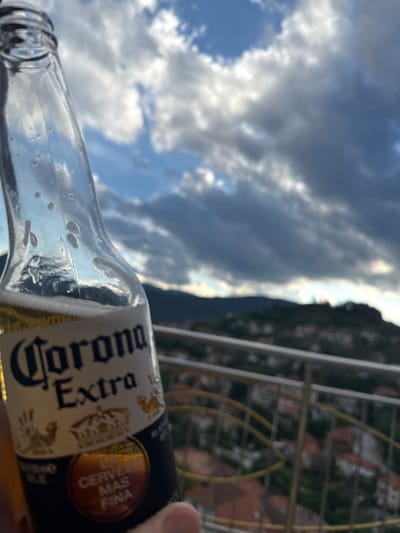 A close-up, shallow depth of field shot captures a glass bottle of Corona Extra beer. The bottle is partially filled with beer and condensation streaks the glass. In the background, a hazy view of a hillside town, likely Momchilivtsi, Bulgaria, is visible under a dramatic, cloudy sky. A metal railing with yellow accents runs horizontally across the lower portion of the frame.