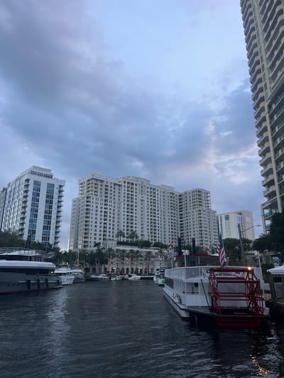 The image depicts a wide canal or river in Fort Lauderdale, Florida, lined with modern high-rise buildings and palm trees. Several boats are docked along the waterway, including a large, white paddlewheel boat with an American flag and smaller yachts. The sky is cloudy, suggesting overcast weather or approaching dusk. The architecture of the buildings is contemporary, featuring balconies and large windows, indicative of a developed urban waterfront.