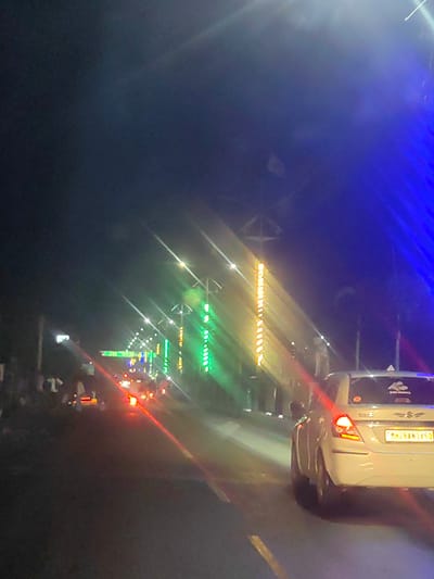 The image captures a nighttime street scene in Mohegaon, India, characterized by a vibrant display of colorful lights. A white sedan is prominent in the foreground, its rear taillights illuminated, and a license plate reading "MH28AN3450" is visible. The car appears to be driving on a road lined with decorative lights, including vertical strings of green, yellow, and white lights. In the background, the silhouettes of people are discernible on the side of the road, suggesting a gathering or event. The overall ambiance is festive and illuminated by artificial lights, typical of a night in an Indian city.