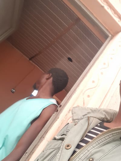 The image depicts a person in Dutsinma A, Nigeria, wearing a light blue tank top and partially obscured by a green jacket with a visible striped shirt underneath. They appear to be standing beneath a light-colored ceiling with recessed lines and a hole. A decorative border runs along the wall or frame. The walls are primarily orange-brown. The perspective is from a low angle, looking upwards at the subjects.