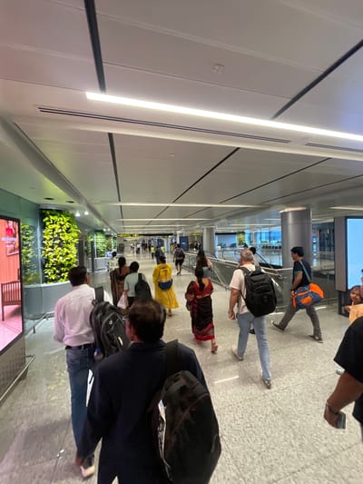 The image depicts a busy interior walkway, likely an airport terminal, with a modern aesthetic. A diverse group of people are walking in various directions, many carrying luggage, suggesting travel. The architecture features a high, illuminated ceiling with long, linear light fixtures and dark structural beams. One side of the walkway is adorned with a large, vertical living wall of lush green plants, adding a natural element to the environment. Several digital screens are visible along the walls, displaying advertisements or information. The flooring is a speckled grey terrazzo. The presence of escalators and directional signage further confirms the setting as a transportation hub. Given the context provided, this scene is situated within the city of Hyderabad, India.