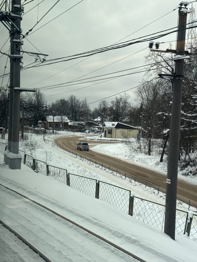 A winter scene shows a road curving through a snow-covered landscape. A car drives along the road, past houses with snow-laden roofs. Power lines stretch across the sky. Fences are also visible, heavily covered in snow. The sky is overcast, creating a subdued, wintry atmosphere.