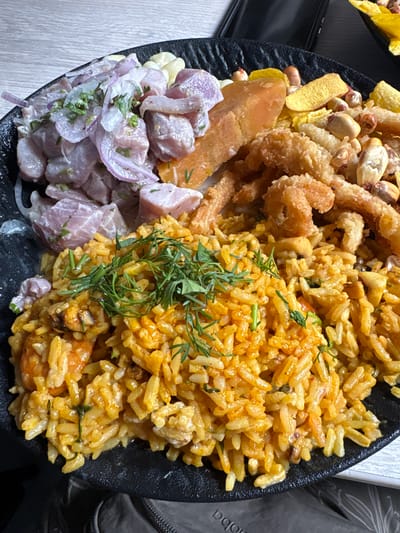 A close-up, overhead shot showcases a diverse platter of Peruvian cuisine on a textured black plate, likely in Jesús María, Peru. The dish is segmented, featuring several distinct preparations.

In the upper left quadrant, crispy plantain chips (chifles), toasted corn (cancha), and peanuts are visible, providing a crunchy texture. Adjacent to this, in the upper center, are pieces of golden-brown fried seafood, possibly calamari or shrimp, indicating a deep-fried preparation.

The main portion of the plate, occupying the right side, is a vibrant yellow-orange rice dish, likely *arroz con mariscos* (rice with seafood), garnished with fresh green herbs resembling dill. Scattered within the rice are chunks of seafood.

On the lower left, a generous serving of *ceviche* is presented, featuring cubes of light-colored raw fish marinated with thinly sliced red onions and visible corn kernels (choclo). A creamy, white dressing or marinade coats the ingredients. Below the fried items and between the ceviche and rice, a soft, orange-hued piece of sweet potato (camote) is placed.

The overall presentation is colorful and abundant, highlighting a combination of popular Peruvian dishes. The plate rests on a light-colored, possibly wooden or laminate, surface. Partially visible on the far right, a gray bag or backpack shows the faint text "poba". The lighting is bright and even, suggesting an indoor setting with good illumination, capturing the textures and colors of the food distinctly. No people are visible; the focus is entirely on the culinary arrangement.