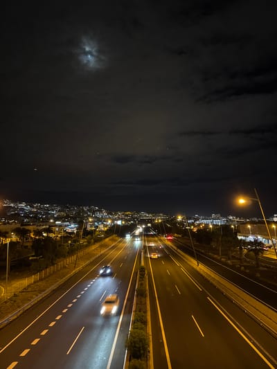 The image depicts a night scene in Adeje, Spain, with a split view showing a city skyline on the right and a cloudy, dark sky on the left. The city is illuminated by numerous lights from buildings and streetlights, creating a vibrant urban landscape. A multi-lane highway runs diagonally through the scene, with cars visible, their headlights and taillights blurred due to motion, suggesting they are in transit. The highway is well-lit by overhead streetlamps, casting a warm glow on the wet asphalt. To the left, the sky is filled with dark, textured clouds, with a faint, diffused light from the moon visible through a gap in the clouds. The overall atmosphere is one of nighttime urban activity contrasted with the atmospheric darkness of the sky.