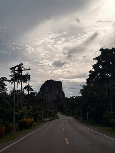 The image shows a road winding through a tropical landscape in Ao Luek, Thailand. The road is paved and marked with a white line and yellow dashes, indicating a two-lane road. On the right side of the road, dense green foliage and trees line the way, with glimpses of lush vegetation. On the left, the sky is filled with dramatic, white, wispy clouds against a lighter gray backdrop, suggesting a partly cloudy or overcast day. In the distance, a large, dark, rocky karsts rises sharply from the landscape, partially obscured by trees. Several utility poles with wires are visible, adding to the rural setting. The overall mood is serene and natural, hinting at a remote or less-developed area. The lighting suggests it could be late afternoon or early morning, with the sun's rays filtering through the clouds. There is no visible text in the image.