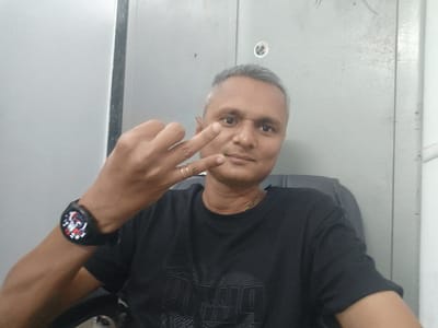 A man is seated in a chair, facing the camera. He is making a hand gesture. He is wearing a black T-shirt and a wristwatch. The background consists of gray metal panels. There are no visible signs indicating the specific location in Dhandhuka, India.