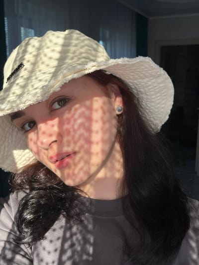 A young woman with dark hair and brown eyes is wearing a light-colored bucket hat and a grey t-shirt. She is looking at the camera with a neutral expression. Sunlight casts a dappled shadow pattern across her face. She has a silver stud earring in her left ear. The background is out of focus and appears to be an indoor setting with a window and curtains. The location is Starobaltachevo, Russia.