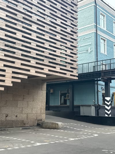 The image shows a street scene in Zhabkino, Russia, with a modern building constructed with wooden slats dominating the left side of the frame. To the right, a blue brick building with white trim and windows is visible, with a metal walkway connecting the two structures at an upper level. The ground level features a dark-colored facade with a grey metallic door and a pillar with black and white chevron markings. The street surface is asphalt with white dashed line markings. No people are present in the image.