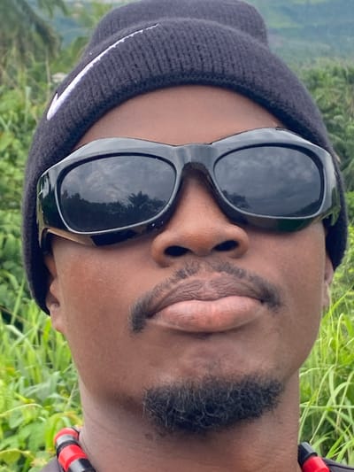 A close-up portrait of a man wearing a black beanie and black sunglasses is presented. The sunglasses reflect a sky with some clouds, potentially suggesting an outdoor setting. The man has dark skin, a mustache, and a beard. The background is blurry, but green vegetation is visible, possibly grass or foliage. The photo is taken at an unusual angle, tilted to the side. The information is limited as to whether the picture was taken in the city of Ufueseni, Nigeria or not.