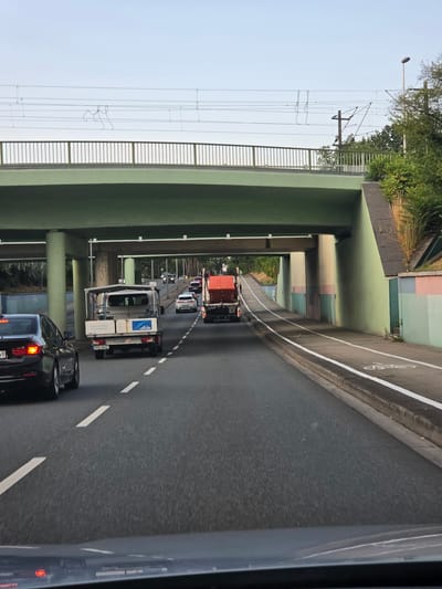 The image depicts a street scene in Rüsselsheim am Main, Germany, viewed from a vehicle driving on a multi-lane road under an overpass. The overpass is painted green and has a metal railing. Overhead, power lines for an electric train are visible. On the road, several vehicles are present, including a black sedan in the left lane, a white utility truck with a canopy and "Linde" branding in the center lane, and a red garbage truck further ahead in the right lane. A separate bike lane is marked with white lines and bicycle symbols along the right side of the road, bordered by a concrete barrier and a painted wall. The environment appears to be urban, with trees and greenery visible beyond the overpass. The time of day seems to be late afternoon or early evening, given the lighting.