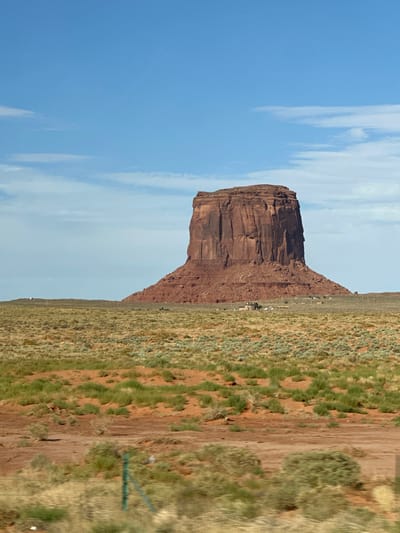 A solitary, massive red sandstone mesa dominates the center of the frame, its sheer cliffs casting shadows across its rugged surface. The mesa rises from a vast, arid landscape characterized by low-lying, scrubby vegetation and patches of reddish-brown earth. A sparse cluster of buildings, possibly homes or outbuildings, is visible at the base of the mesa. The sky above is a clear, vibrant blue, with a few wispy white clouds scattered across it. The foreground of the image is blurred, suggesting the photo was taken from a moving vehicle, and a green metal fence post is visible on the left side of the frame. The scene depicts a characteristic vista of Oljato-Monument Valley, United States of America.