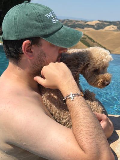 A person wearing a green baseball cap and a bracelet is relaxing by a pool with a fluffy, light brown dog. The cap has white text that reads "THE ISSI." The person's arm, which is hairy, rests on the dog, and their hand is near the dog's head. The dog appears to be a poodle mix. The scene is outdoors with a bright, sunny sky, suggesting it is daytime. The background shows the blurred blue water of a swimming pool and a sun-drenched landscape with rolling hills and dry vegetation, consistent with a Mediterranean climate like Asciano, Italy. The overall mood seems relaxed and peaceful, with the person enjoying the company of their dog in a warm, outdoor setting.