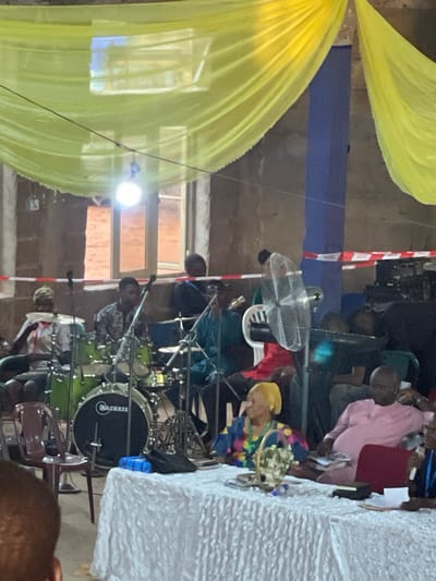 The image depicts an indoor setting, possibly a church or event hall, in Nteje, Nigeria. A green drum set is positioned in the foreground, with drummers partially visible. A white-covered table is set to the right. Yellow fabric drapes from the ceiling, indicating a decorated space. A group of people is gathered behind the drums, with clothing colors including pink, green, and various patterns. Other objects include a standing fan, microphone stands, and chairs. The walls and pillars appear to be unfinished or minimally decorated. There are windows and electric wiring visible on the walls.
