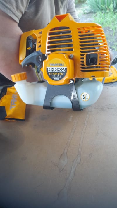 A top-down, medium shot captures a person in a light-colored shirt holding a bright orange gasoline brush cutter. The brush cutter is the primary subject, with its fuel tank, engine housing, and control handle clearly visible. A yellow fabric accessory, possibly a carrying bag or cover, is positioned to the left of the brush cutter. The background shows a blurred natural setting with green foliage, suggesting an outdoor environment. The brush cutter is placed on a textured, light brown surface, which appears to be a table or workbench. A label on the brush cutter reads "TAHATSU BENZOKOSA GASOLINE BRUSH CUTTER TS-43R PRO." A circular yellow sticker with a graphic is also visible on the fuel tank. The lighting is diffused, and the overall image has a slightly muted color palette. The scene appears to be taking place in or around Bagayevskaya, Russia, likely in a residential or workshop setting where such equipment would be stored or maintained.