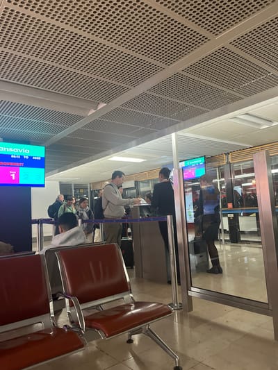 The image shows the boarding area in what appears to be an airport in Paray-Vieille-Poste, France, based on the airline branding of Transavia, which operates flights from Paris-Orly Airport located in that city. There is a monitor displaying flight information, showing gates numbered 1, 2, and 3. People are standing in line at the gates, presumably waiting to board their flight. There are rows of empty chairs, mostly brown leather or leatherette, in the foreground. The background has architectural elements of the building including the ceiling design, which is perforated metal. A gate counter with a glass partition separates passengers from staff. A woman stands at the gate.