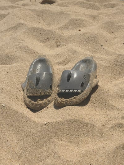 The image shows two gray shark-shaped sandals partially buried in sand. The location is a sandy beach, likely in Puerto Escondido, Mexico. The sandals are oriented with the 'shark faces' facing upwards. The sand is light brown and covers portions of the sandals, suggesting they have been there for some time. No people are visible. The scene consists of discarded sandals on a beach.