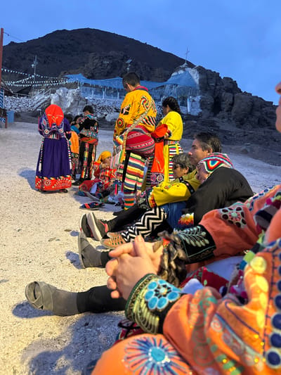 A group of people wearing colorful traditional clothing are standing together. The outfits are predominantly orange, yellow, and blue with intricate embroidered designs. The people are close together, suggesting a sense of community. In the background, there's a mountain, a small white building with a cross on top, and festive decorations, indicating a celebration or cultural event. The scene is bathed in the soft light of dusk or dawn.