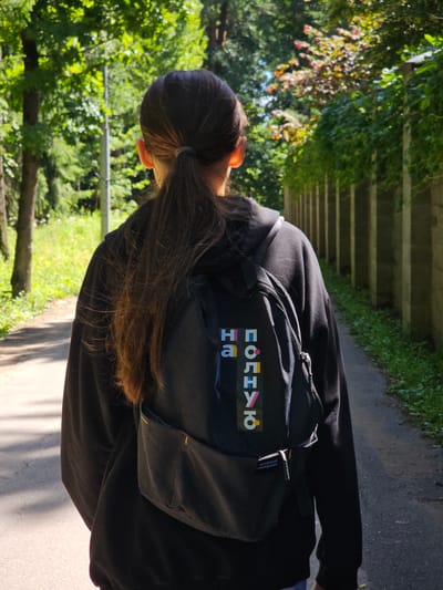 A young person with long, dark hair pulled into a ponytail, wearing a black hooded sweatshirt and a black backpack, is walking away from the camera on a paved path. The backpack has a colorful patch with Cyrillic text that reads "НА ПОЛНУЮ". The path is bordered by green grass and trees on the left and a tall concrete fence on the right. The scene appears to be a park or a quiet street in a residential area. The lighting suggests it is daytime with sunlight filtering through the trees.