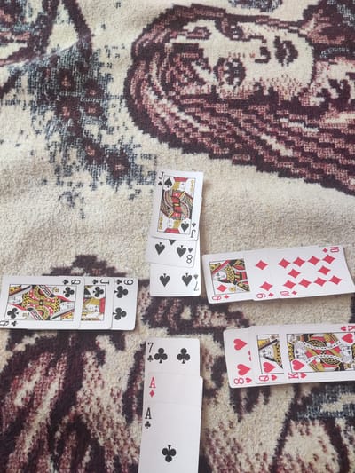 This image depicts a close-up, overhead view of playing cards laid out on a patterned rug. The rug has a creamy white base with abstract, dark red and brown patterns.
The playing cards visible are:
* A stack of three cards, face up, showing the Jack of Spades at the top, followed by an 8 of Spades, and another card partially obscured.
* A row of three cards, face up, showing the Queen of Clubs, a Jack of Clubs, and a 9 of Clubs.
* A row of two cards, face up, showing the Queen of Diamonds and the 10 of Diamonds.
* A row of three cards, face up, showing the 8 of Hearts, the Queen of Hearts, and the King of Hearts.
* A stack of three cards, face up, showing the 7 of Clubs at the top, followed by an Ace of Diamonds, and an Ace of Clubs.
The lighting suggests an indoor setting, possibly in a home. There are no people visible in the image. The specific location of Manole, Bulgaria is not discernible from the visual content of the image itself.