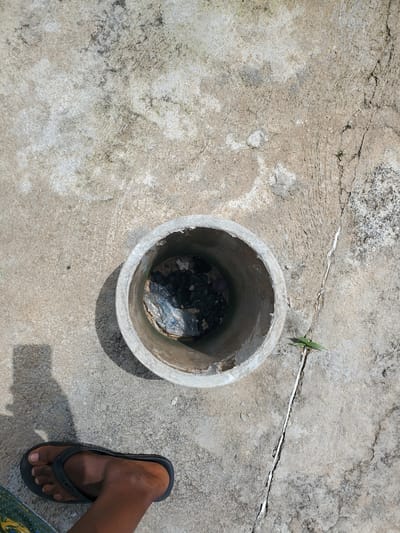 An overhead shot shows a person's bare left foot wearing a black flip-flop standing on a concrete surface. To the right of the foot is a circular, concrete pipe with its opening facing upwards. Inside the pipe are dark-colored plastic bags and what appears to be debris. The concrete surface is weathered and cracked in several places, with a thin vine growing through one of the larger cracks. A shadow from the pipe is cast on the concrete to the left. The scene is lit by harsh sunlight, creating strong contrasts. The location is identified as Kason, Nigeria.