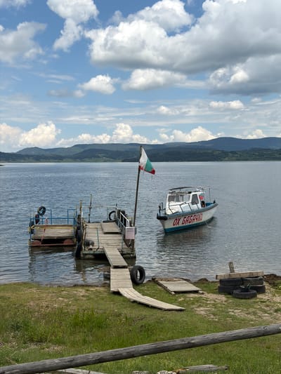 The image depicts a tranquil lakeside scene in Цигов чарк, Bulgaria, on a day characterized by a blue sky with numerous large, white cumulus clouds. In the background, a range of dark green, forested mountains stretches across the horizon, meeting a calm, grey-blue body of water, likely Batak Lake.
In the midground, a rustic wooden dock extends into the water from the grassy shore. The dock consists of multiple interconnected floating wooden platforms, equipped with rudimentary metal railings. Several old tires are integrated into the dock structure, serving as fenders or flotation. A wooden pole on the main dock section proudly flies a Bulgarian flag, displaying horizontal white, green, and red stripes. A small pink sign on the dock bears Cyrillic text: "РАЗХОДКИ (088 850 70)", which translates to "WALKS (088 850 70)", indicating boat excursions are offered, along with a contact number.
To the right of the dock, a white motorboat with a blue stripe on its hull is moored in the water. The boat has a small cabin, and red Cyrillic lettering on its side reads "ОХ БАБАЧКО". A small Bulgarian flag is also visible on the boat. A faint figure, possibly a child, can be seen inside the boat's cabin through a window.
The foreground consists of a grassy bank leading to the water's edge. Multiple wooden plank walkways connect the shore to the floating dock. On the grassy area to the right of the dock, a stack of old tires is present, along with a single tire lying flat. A rough, weathered wooden log or fence runs horizontally across the very bottom of the frame, partially obscured by grass. The water surface shows minor ripples, reflecting the sky and distant features.