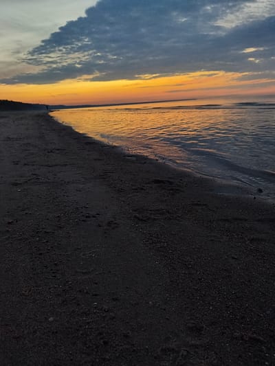 The image depicts a serene beach scene at dusk in Apšuciems, Latvia. The foreground is dominated by dark, wet sand with visible footprints and small pebbles. The gentle waves of the Baltic Sea lap at the shore, reflecting the vibrant colors of the sunset. The horizon is a band of warm orange and yellow hues, transitioning into a pale pink and then a dusky blue as it meets the sky. Scattered, dark clouds drift across the upper portion of the sky, adding depth and texture to the twilight atmosphere. A lone figure is faintly visible walking along the shoreline in the distance, a silhouette against the illuminated sky. The overall impression is one of tranquility and the natural beauty of the Latvian coast during sunset.