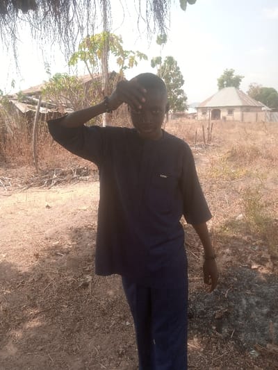A young male stands centrally in a dry, open area within Gudige Sabo, Nigeria. He is dressed in a dark blue short-sleeved top featuring a left chest pocket and dark blue trousers. Dark beaded bracelets are visible on both wrists. His left hand is raised to his forehead, fingers splayed across his brow, as he looks directly into the camera. The ground beneath him is dry, reddish-brown earth, sparsely covered with withered grass, dry leaves, and small scattered debris. In the background, to the left, a structure with a partially damaged or collapsed corrugated metal roof is visible. To the right, a single-story building with light-colored walls and a conical corrugated metal roof stands. Sparse green foliage from trees and bushes is distributed throughout the background. The sky is bright and clear.