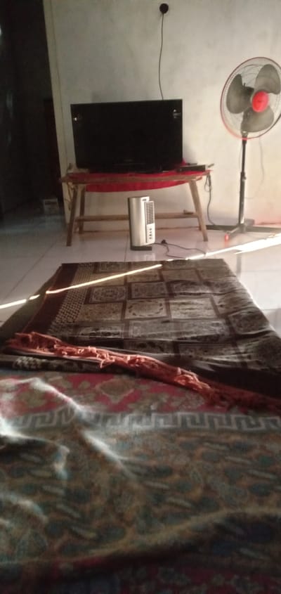 This indoor scene appears to be in a home located in Bulukandang, Indonesia, featuring a living room-like setup. A flat-screen television sits on a wooden stand against a white wall, with a tower fan placed to its right. A portable space heater is located under the stand. A patterned rug covers a significant portion of the floor, with a decorative runner or scarf draped across it. The room is brightly lit by sunlight streaming through a window, creating a strong contrast of light and shadow. The floor appears to be tiled.
