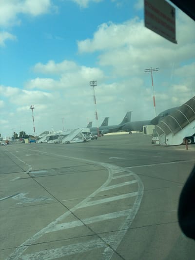 The image depicts an airport tarmac on a cloudy day, with the sky a brilliant blue with white cumulus clouds. In the foreground, the concrete surface of the tarmac is marked with white painted lines, including directional arrows and a large curved pattern. To the right, a portion of a black object, possibly a vehicle's interior or a person's head, is visible.
Further back on the tarmac, several aircraft are parked. The most prominent is a large, dark-colored jet with a distinctive hump-like structure on its fuselage, suggesting it may be a military tanker aircraft. Two other similar aircraft are parked behind it, also with their tails visible.
Adjacent to the aircraft are mobile staircases, some of which are connected to the planes, indicating passengers or crew may be boarding or disembarking. A white bus with "COBUS" written on its side is positioned near one of the staircases. Also visible are support vehicles and lights on tall poles with red and white striped markings, common airport navigational or lighting towers.
In the distance, a line of trees and some buildings can be seen, hinting at the airport's surroundings. The overall scene is one of activity at an airport, with aircraft, ground support equipment, and infrastructure present. The location is identified as Yagel, Israel, suggesting this is an airport in that region.