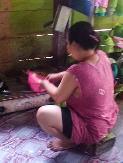 In Pompanua, Indonesia, a woman is crouched on a low stool in front of a stove, cooking. She is wearing a pink t-shirt and black shorts. Her hair is pulled back in a bun. A pink bowl sits on the stove next to a pot. The environment appears to be an outdoor kitchen, with wooden walls and a tiled floor. Various green containers are visible on shelves in the background, as well as a blue container and some pink cups.