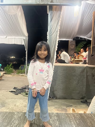 A young girl, appearing to be of Indonesian descent and dressed in a long-sleeved pink and white shirt with cartoon graphics and blue jeans, stands in the foreground of an outdoor scene. She is facing the camera with a slight smile, her hands clasped in front of her. Behind her, a group of men are seated on a raised platform, some of whom appear to be playing musical instruments, possibly drums. The area is decorated with white draping fabric, and there are speakers visible. The ground is a concrete surface with several pairs of sandals scattered about. The background suggests a rural or semi-rural setting with some vegetation and structures visible in the dim light. The overall ambiance suggests an informal gathering or performance, possibly a cultural event, taking place in the evening. The image is taken in Wedarijaksa, Indonesia, a sub-district in the Pati Regency of Central Java.