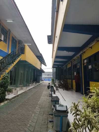 This image depicts an outdoor walkway between two school buildings. The buildings are two stories high, painted in bright yellow and green with blue accents. The walkway is paved with gray bricks. On the left side of the walkway, there are several white cube-shaped benches and a lush green plant. On the right side, there is a covered walkway with several tables and chairs. Further down the walkway, there are multiple blue and gray trash cans. In the background, a group of people wearing blue uniforms are gathered. A person in a maroon shirt stands near a doorway. The sky is overcast. This scene is located in Katrungan, Indonesia, likely within a school campus due to the uniforms and architecture.