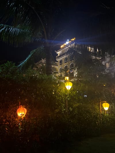 The image depicts a nighttime scene in Hoi An, Vietnam, characterized by warm, ambient lighting and lush greenery. The foreground is dominated by the fronds of a palm tree, their dark silhouettes contrasting with the illuminated foliage and a multi-story building in the background. The building appears to be a hotel or resort, with arched windows and decorative architecture visible beneath the illumination.

Several decorative lanterns, shaped like stylized hot air balloons, are strategically placed throughout the scene. These lanterns emit a soft, yellow-orange glow, illuminating the surrounding bushes and trees. One lantern is particularly prominent in the lower right quadrant of the image, casting a warm light on the dense vegetation. Another lantern, higher up and to the left, also contributes to the overall ambiance. A blue light ray streaks diagonally from the upper left, possibly a lens flare or light from a distant source.

The scene suggests a peaceful and inviting atmosphere, typical of a tropical evening in a tourist destination. The time of day is clearly night, as indicated by the dark sky and artificial lighting. The lush vegetation hints at a warm climate, and the architectural style of the building may suggest a colonial influence, common in historical Vietnamese cities like Hoi An.

Visible text on the building, partially obscured by the palm tree, reads "NES VA". The overall impression is one of a serene and picturesque setting, perfect for an evening stroll or relaxation.