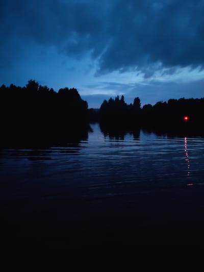 The image depicts a dark, serene river scene at dusk, situated in the vicinity of Badozhskoi Pogost, Russia. The sky is a deep, stormy blue, with heavy clouds casting a somber mood. Silhouetted against the sky is a dense line of trees forming the riverbank, their forms indistinct in the low light. The river itself is calm, with gentle ripples disturbing its dark surface, reflecting the muted tones of the sky. In the middle distance, a solitary red light, possibly a buoy or marker, emits a faint glow, its reflection stretching down into the water in a series of shimmering red dashes. There are no people visible in the frame, and the overall impression is one of quiet solitude and natural beauty. The image captures a moment of transition, as day gives way to night, with the natural elements taking center stage.