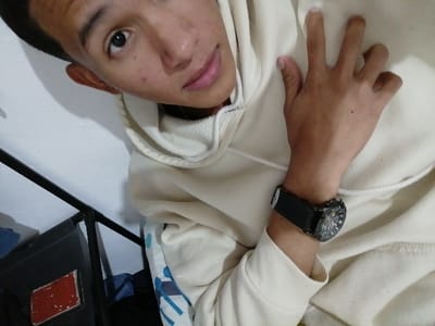 A person is pictured wearing a beige hoodie and a black wristwatch. Their face is visible, along with one hand partially obscuring their chest. The background includes a white wall and a glimpse of a black metal structure with some items nearby. The overall perspective is from above, looking down on the subject.
