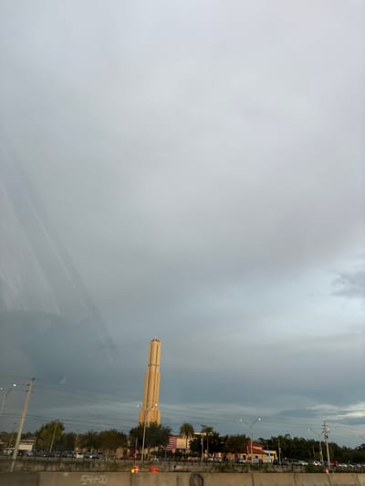 The image captures a view of the city of Sarasota, Florida, United States, under a cloudy sky. In the foreground, a concrete barrier with graffiti is visible, along with a tire and orange traffic cones. Beyond the barrier, a highway with multiple lanes of traffic is present, indicated by cars and a traffic light.
Dominating the middle ground is a tall, slender tower with a distinctive stepped design and a pointed top. The tower is illuminated by lights, casting a warm glow against the overcast sky. Adjacent to the tower are various buildings, some with red roofs, and numerous trees. An American flag is prominently displayed on the side of one of the buildings.
The background consists of a vast expanse of gray, cloudy sky. The overall scene suggests an urban environment, possibly near a commercial or recreational area, under a dramatic or impending weather condition. The lighting and cloud cover create a slightly somber or atmospheric mood.