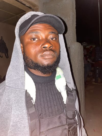 The image shows a medium shot of a Black man wearing a gray hooded jacket, a black baseball cap, and a black turtleneck sweater. He has a beard and is looking directly at the camera. He is also wearing what appears to be a tactical vest with straps and D-rings. The background is out of focus but appears to be an outdoor environment at night, possibly a street or an open area with some structures and people in the distance. The lighting suggests artificial light sources.
Based on the provided information, it is not possible to determine the specific location within the city of Jos, Nigeria, as no identifying landmarks or contextual clues are present in the image. The image itself does not contain any specific details that can pinpoint its location to Jos.