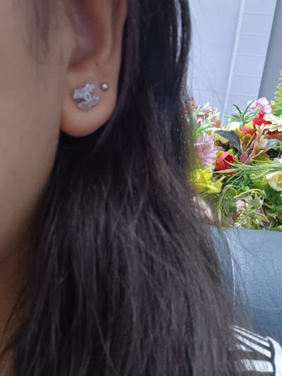 In Shahand, Uzbekistan, a close-up shot focuses on a person's ear, adorned with a small, silver stud earring and a prominent Chanel logo stud earring. The person's dark, long hair cascades down, partially obscuring the background. To the right, a vibrant bouquet of artificial flowers, featuring shades of pink, red, yellow, and green, adds a splash of color. The background appears to be an indoor setting with a textured wall and a faint pattern on a surface, possibly a couch or chair.