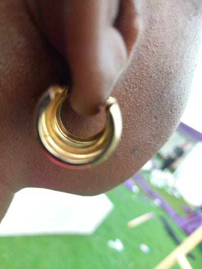 An extreme close-up depicts a person's right earlobe, showcasing brown skin with visible texture. A gold-toned, multi-layered hoop earring is worn in the earlobe piercing. The background is out of focus, revealing a green surface consistent with artificial turf or grass, upon which indistinct white objects are situated. Vague purple and yellow structures are discernible in the blurred far background, suggesting outdoor equipment. The specific location within Ohafia, Nigeria, cannot be ascertained from the visual evidence presented.