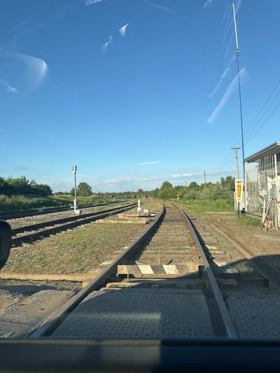The image shows a view of a railway crossing on a clear day. Multiple railway tracks run parallel to each other, curving to the right in the distance. Green vegetation and trees are visible on either side of the tracks, with a bright yellow sign with Russian text that reads "ПОСТ" (POST) placed near a building on the right side of the image. A tall pole with some equipment is also present near the sign. The foreground shows the surface of a road or pathway at the crossing, made of concrete and metal plates. The sky is clear blue with a few wispy white clouds. The scene appears to be in an outdoor, rural or semi-rural setting, likely within the vicinity of Кварса, Russia, given the presence of Russian text on the sign and the overall landscape. There are no people or active vehicles visible in the frame.