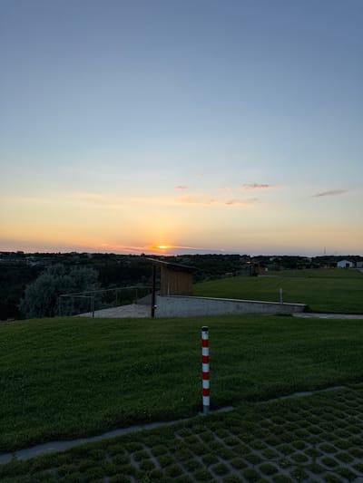 A low-angle, full outdoor shot depicts a scenic view of Topola, Bulgaria at sunset. The sky is a gradient of light blue and orange, with the sun partially obscured by a strip of clouds near the horizon. Below the sky, a lush green landscape stretches out, dotted with trees and buildings. In the foreground, a red and white striped pole stands upright on the grass. A pathway of permeable paving stones is visible in the bottom right corner. The setting appears to be a park or recreational area, with several small, wooden structures scattered across the grassy fields. The overall impression is one of tranquility and natural beauty.