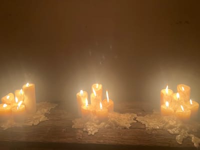 Illuminated by candlelight, a dimly lit scene features three clusters of varying sized, partially burnt candles sitting atop a rustic, wooden surface. An abundance of melted wax has accumulated beneath each cluster, solidifying into irregular shapes. The background is a blurry, neutral tone, possibly a wall. A hazy, smoke-like effect obscures parts of the image, contributing to an atmospheric, almost ethereal, aesthetic.